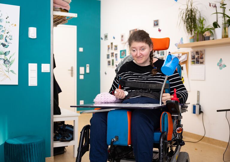 Eine Kundin im Rollstuhl sitzt in ihrem Zimmer und lächelt fröhlich in die Kamera.