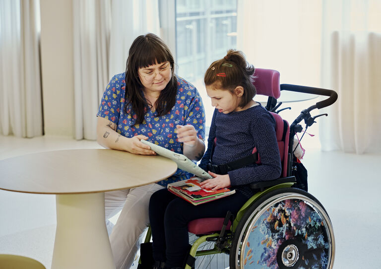 Mitarbeiterin sitzt mit junger Bewohnerin im Rollstuhl beim Tisch und zeigt ihr etwas auf der Tafel Mitarbeiterin sitzt mit junger Bewohnerin im Rollstuhl beim Tisch und zeigt ihr etwas auf der Tafel