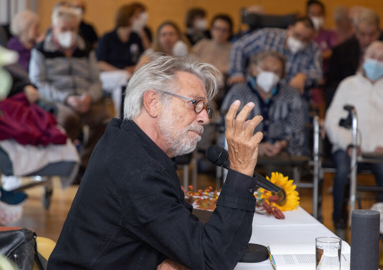 Adi Hirschal seitlich, hinter ihm unscharf die Bewohner*innen.