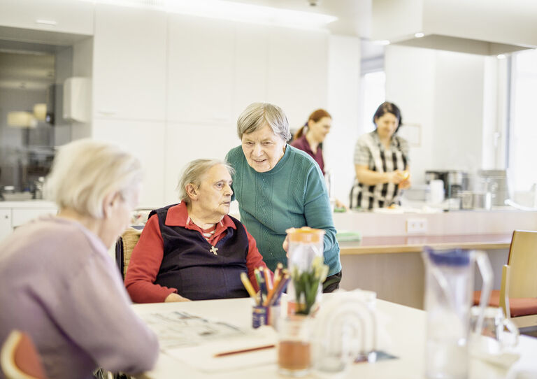 Drei Bewohnerinnen unterhalten sich am Esstisch, im Hintergrund wird das Essen zubereitet.