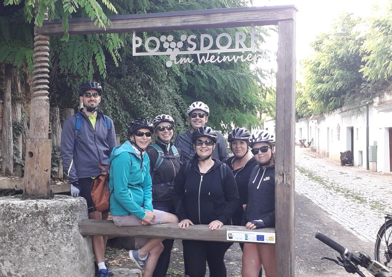 Die Mitarbeiter*innen stehen im Fahrradoutfit für ein kleines Gruppenbild zusammen. Sie befinden sich ein einem Outdoor-Bilderrahmen mit dem Schriftzug "Poysdorf Vinoversum"