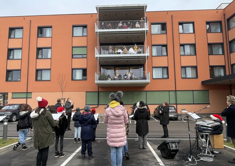 einige Schüler*innen stehen am Parkplatz und singen unter der Leitung ihrer Lehrer*innen. Im Hintergrund stehen auf vier Stockwerken die Bewohner*innen am Balkon und hören zu.