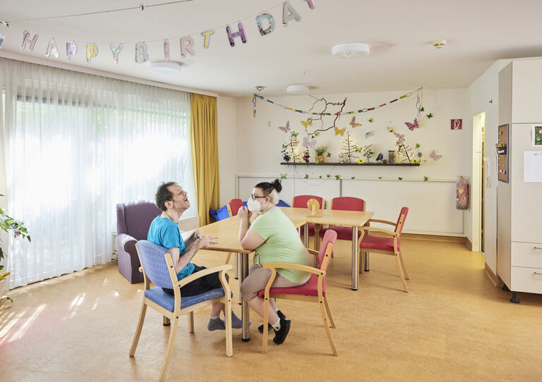 ein Kunde und eine Mitarbeiterin sitzen in der Wohnküche der HABIT WG Attemsgasse bei Tisch und kommunizieren miteinander.