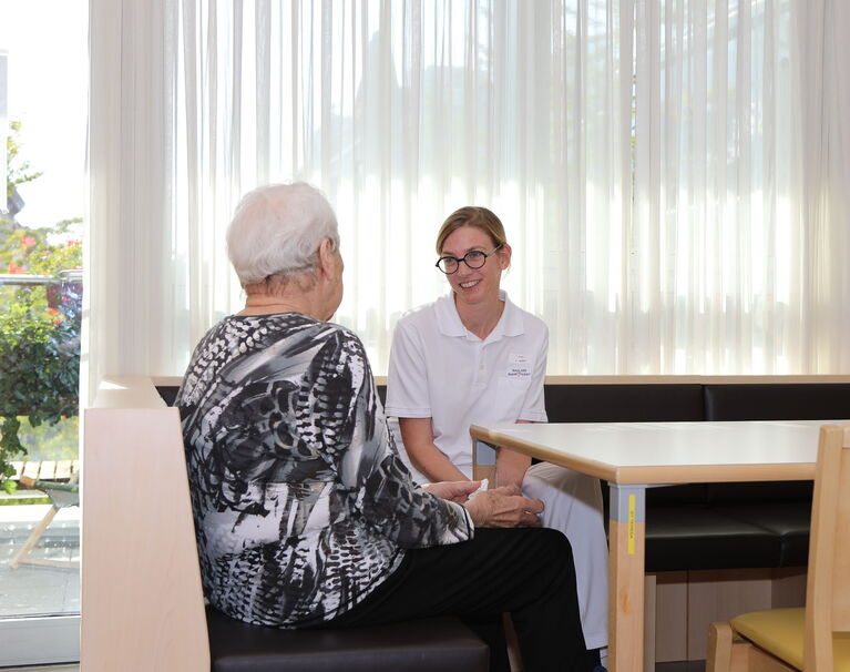 Gespräch mit Ärztin Bewohnerin sitzt Pflegemitarbeiterin an einem Tisch gegenüber. Bewohnerin sieht man von hinten. Die Pflegerin von vorne.