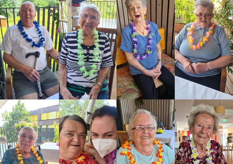 Acht Bewohner*innen und eine Mitarbeiterin lächeln fröhlich und mit einer Blumenkette behängt in die Kamera.