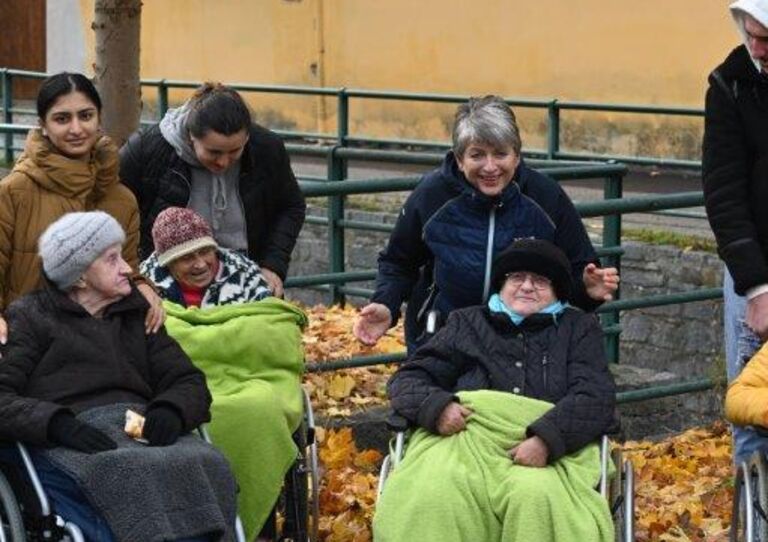 Bewohner*innen im Rollstuhl mit Decke unter einem Baum. Hinter dem Rollstuhl stehen die Mitarbeiter*innen.