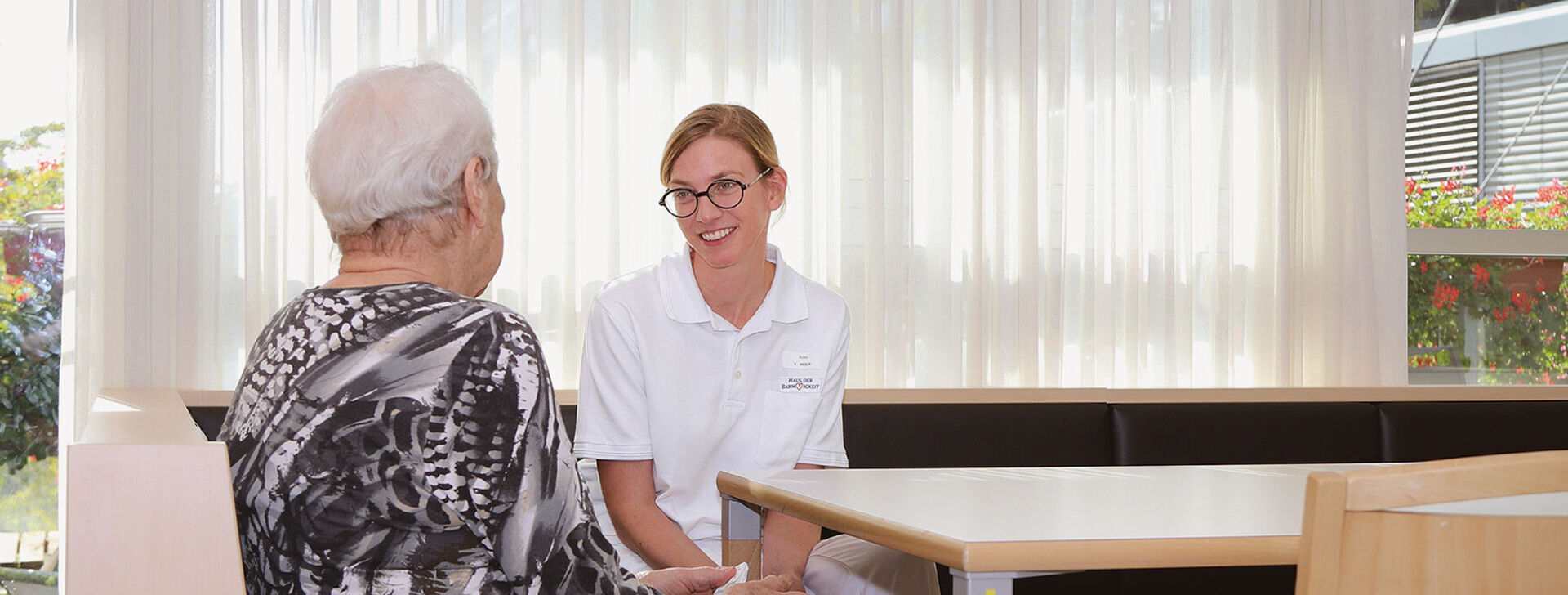 Bewohnerin sitzt Pflegemitarbeiterin an einem Tisch gegenüber. Bewohnerin sieht man von hinten. Die Pflegerin von vorne.