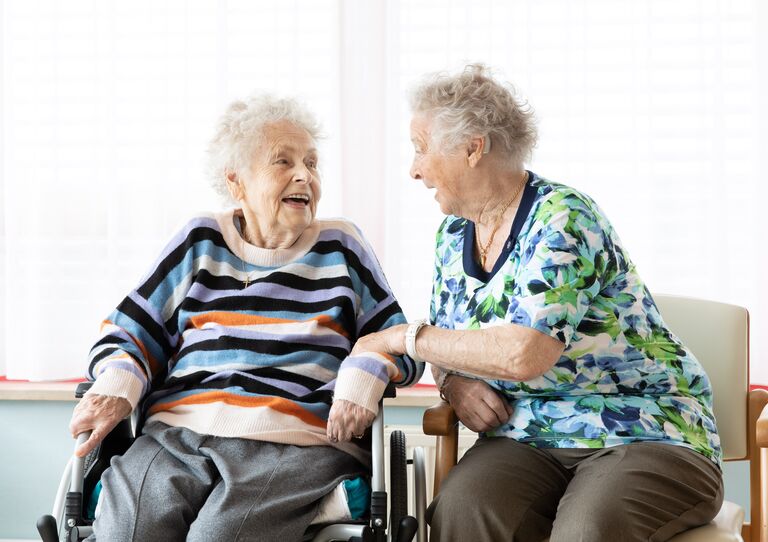 Zwei Bewohner*innen sitzen nebeneinander, unterhalten sich und lachen miteinander. Eine Bewohnerin berührt den Arm der anderen.