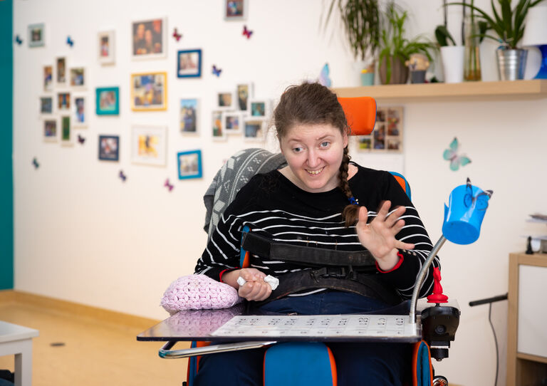 Eine Kundin im Rollstuhl, sie winkt in die Kamera.