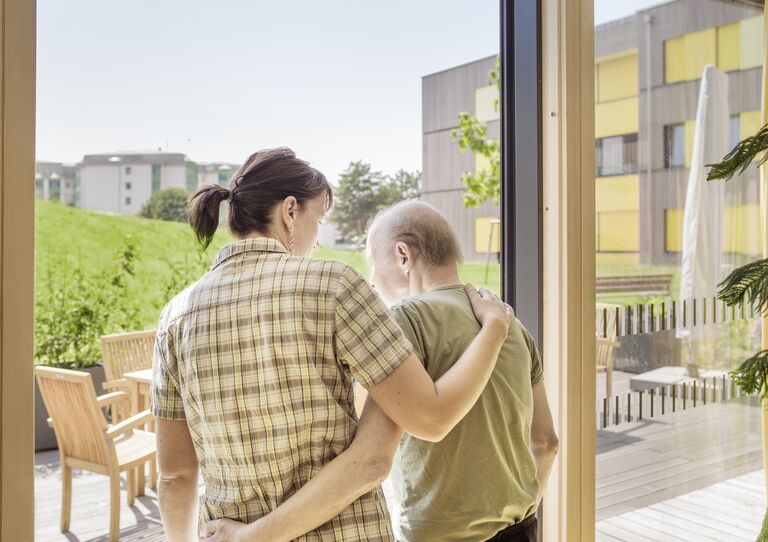 Mitarbeiterin stützt und umarmt Bewohner von hinten - beide gehen auf den Balkon Mitarbeiterin stützt und umarmt Bewohner von hinten - beide gehen auf den Balkon