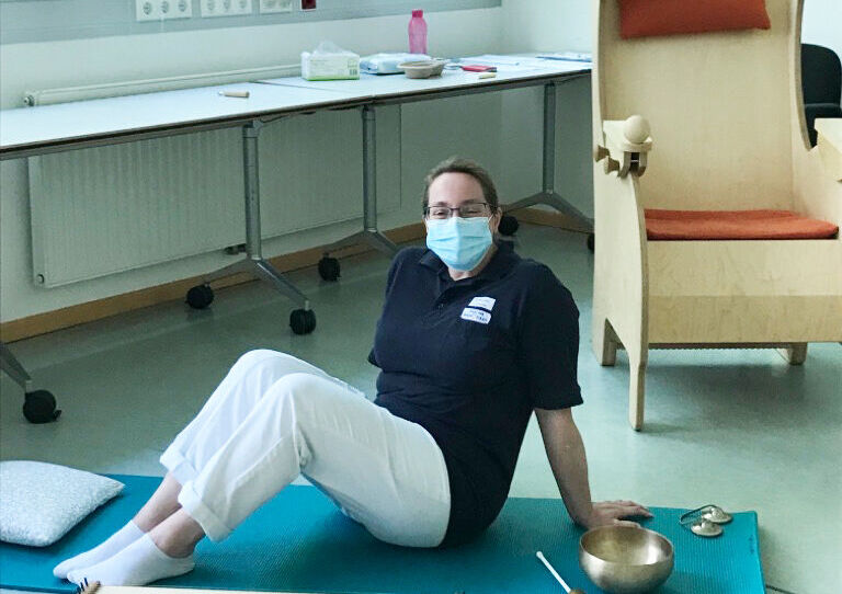 Eine HB Mitarbeiterin sitzt auf der Matte und rund um sie herum finden sich verschiedene Instrumente für die Musiktherapie.