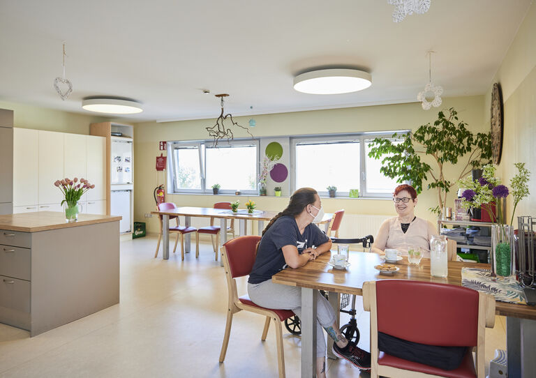 Im Wohn- und Speisezimmer der HABIT WG Arndtstraße sitzen eine Kundin und eine Mitarbeiterin beim Tisch und unterhalten sich