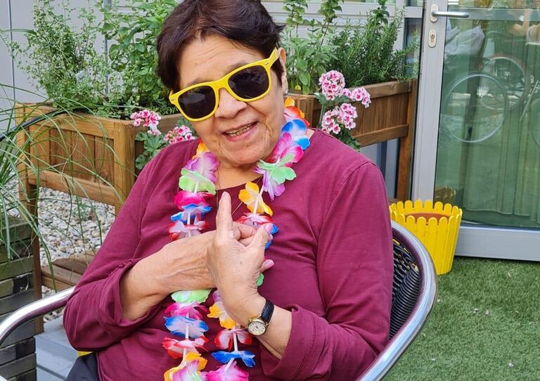 Bewohnerin sitzend klatscht in die Hände und trägt eine Sonnenbrille und Blumenkette
