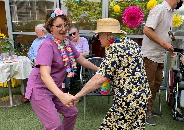 Mitarbeiterin tanzt mit einer Bewohnerin, die einen Sonnenhut trägt.