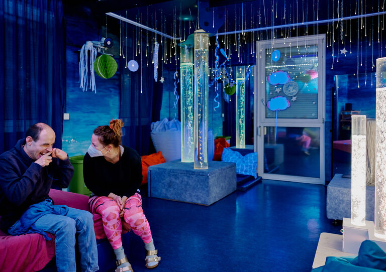 BTZ Lobenhauerngasse: Snoezelenraum: eine Mitarbeiterin sitzt neben einem Kunden, der lächelt. 