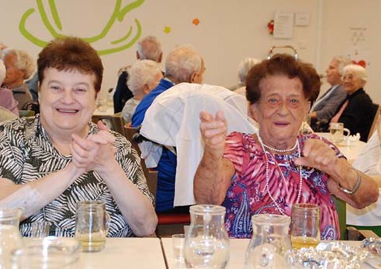 Zwei Bewohnerinnen sitzen nebeneinander und klatschen in die Hände. Das Foto wurde im Rahmen eines Oktoberfests aufgenommen.