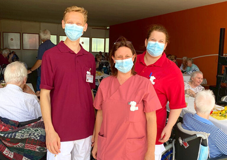 Drei Mitarbeiter*innen aus dem Clementinum lachen in die Kamera hinter ihnen sitzen die Bewohner*innen auf zwei Tischen