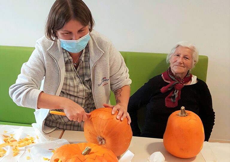 Eine Mitarbeiterin höhlt einen Kürbis aus, eine Bewohnerin schaut dabei zu.
