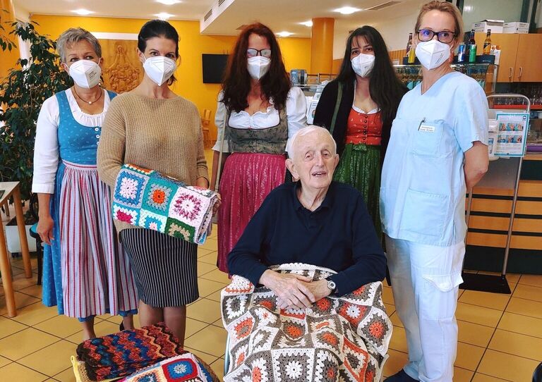 Bewohner im Rollstuhl mit selbst gehäkelter Decke, hinter ihm Wollweiber und Kolleg*innen aus dem HB Clementinum, die teilweise weitere Decken in der Hand halten.