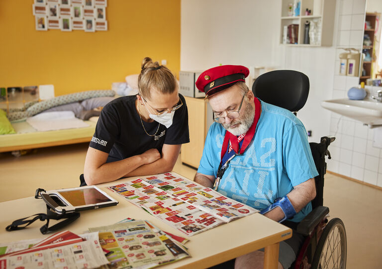 Ein Mitarbeiter sitzt neben einem Kunden im Rollstuhl und sieht mit ihm ein Prospekt an. Der Kunde trägt eine Bahnmütze.