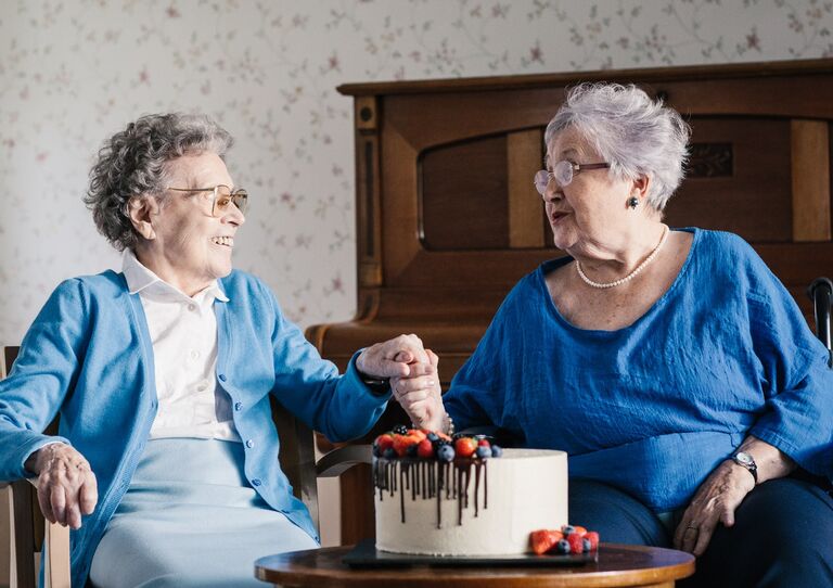Zwei Bewohnerinnen sitzen nebeneinander, halten sich an den Händen und vor ihnen steht eine Torte.