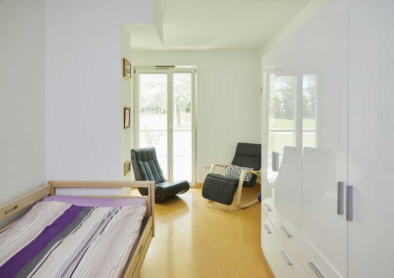 Individuelles Kund*innen-Zimmer der HABIT WG Thürnlhofstraße mit Bett und weißem, glänzenden Schrank und zwei Schaukelstühlen am Fenster 