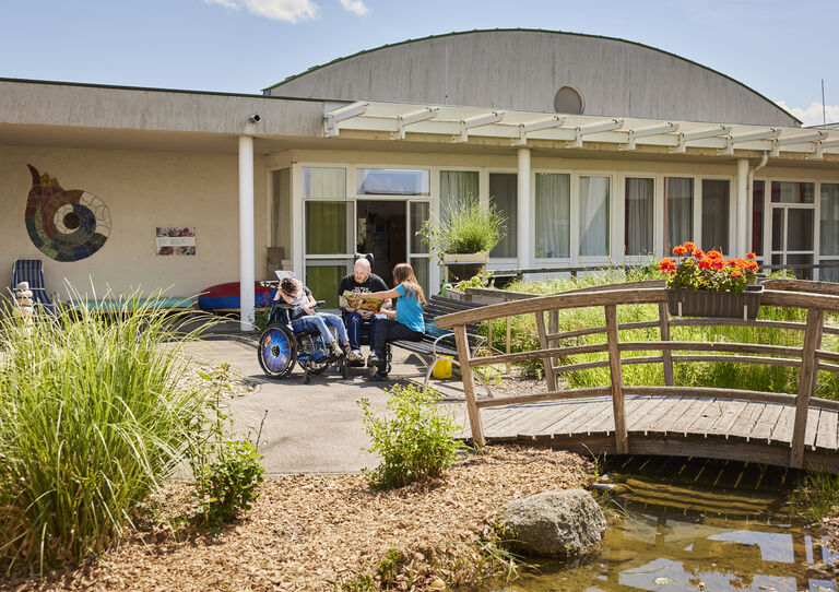 Aufnahme vom Garten. Eine Mitarbeiterin sitzt neben zwei Kundinnen. Gemeinsam schauen sie etwas an. 