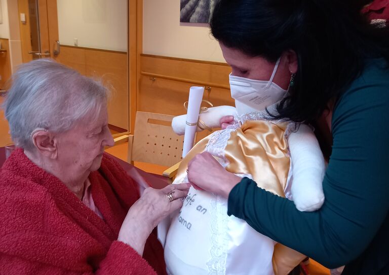 Bewohnerin Elsa schaut eine kleine Schutzengelpuppe an, die ihr Mitarbeiterin Daniela überreicht.