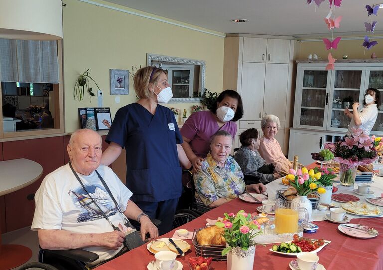 Mitarbeiter*innen und Bewohner*innen posieren für ein Gruppenbild. Am Tisch steht ein reichlich gefüllter Esstisch mit Frühstück.