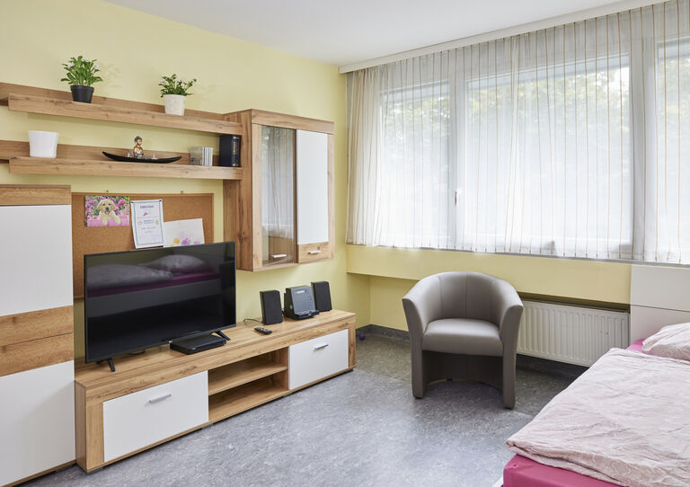 Kund*innenzimmer der HABIT WG Arndtstraße mit Wohnwand mit Fernseher und Fauteuil am Fenster