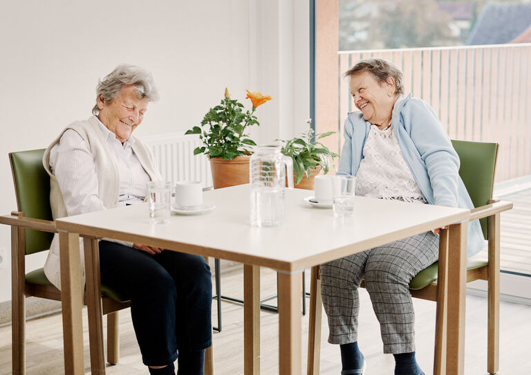 Zwei Bewohnerinnen aus dem Stadtheim sitzen lachend nebeneinander auf einem Tisch