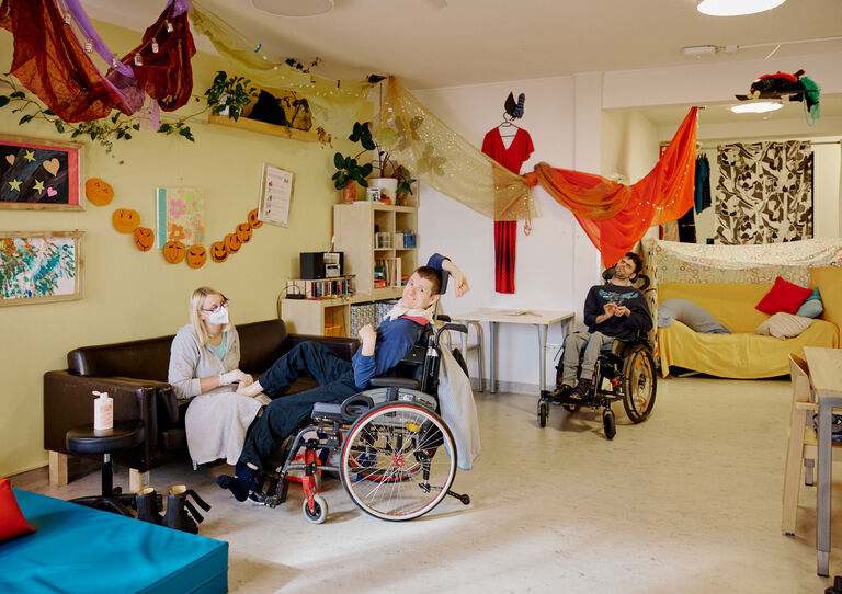 BTZ Lobenhauerngasse: Eine Mitarbeiterin sitzt auf der Couch, ein Kunde sitzt ihr im Rollstuhl gegenüber und streckt seinen Kopf zurück in Richtung Kamera. 