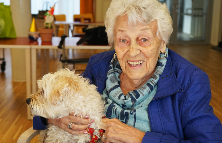 Die Bewohnerin sitzt beim Tisch und hält den Hund auf ihrem Schoß.