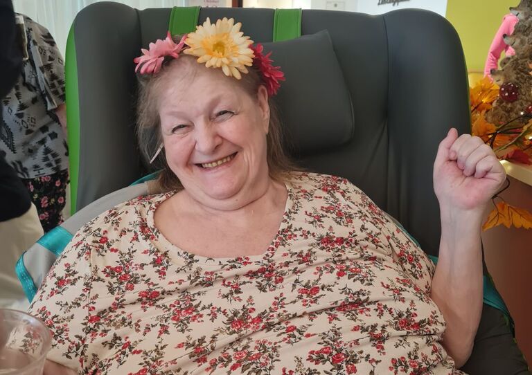 Eine Bewohnerin mit Blumenkranz sitz im Sessel und lächelt in die Kamera.