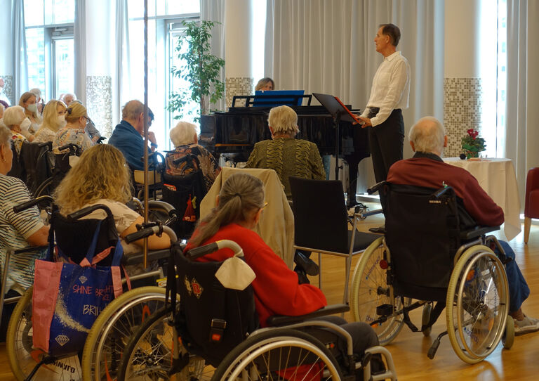 René Rumpold singt und streckt die Hand in die Höhe, links daneben steht ein Klavier und davor sitzt das Publikum.