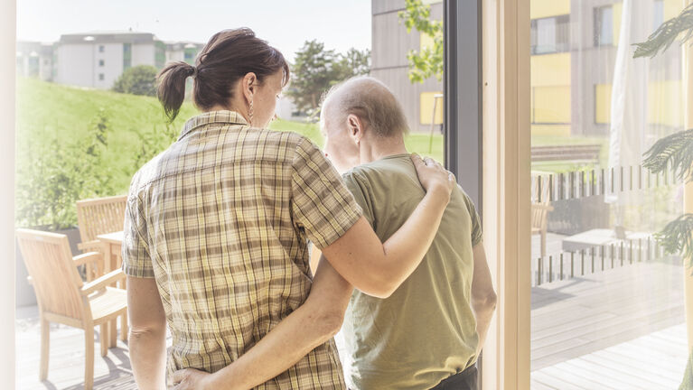 Mitarbeiterin legt Arm um Hospiz-Bewohner. Beide sind von hinten zu sehen.