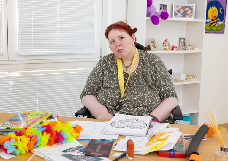 Eine Kundin im Rollstuhl sitzt an ihrem Schreibtisch. Vor ihr liegen viele Bilder und Bastelutensilien.