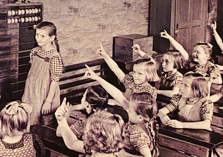 Schwarz-Weiß Foto einer Schulklasse von früher - alle Kinder heben die Hand, ein Mädchen steht auf der Seite und trägt zwei Zöpfe