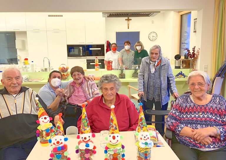 Bewohnerinnen sitzen rund um den Tisch und lachen in die Kamera, vor ihnen die selbstgebastelten Faschingspuppen, im Hintergrund drei Mitarbeiterinnen.