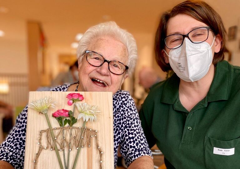 Bewohnerin hält Bild mit Blumenvase hoch, Mitarbeiterin sitzt neben ihr.