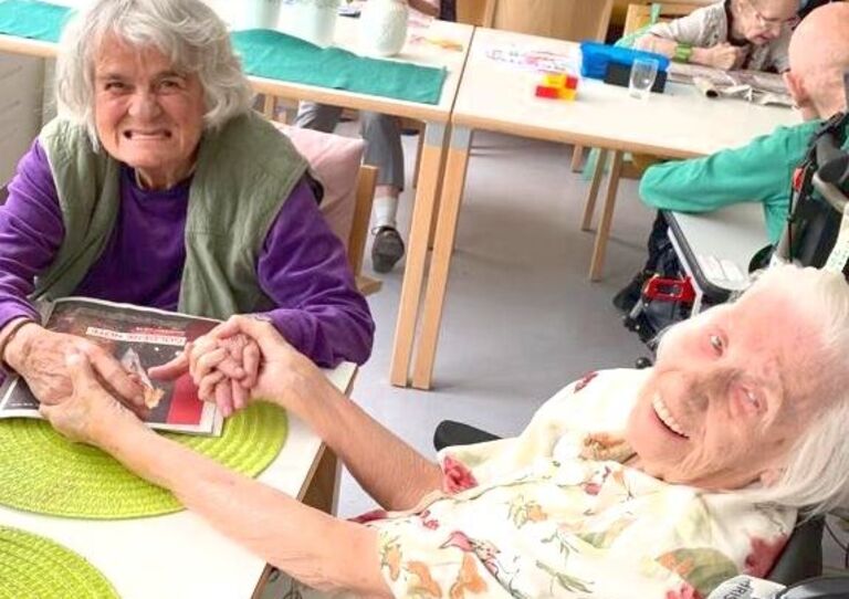 Zwei Bewohner*innen sitzen beim Tisch und halten einander die Hände.