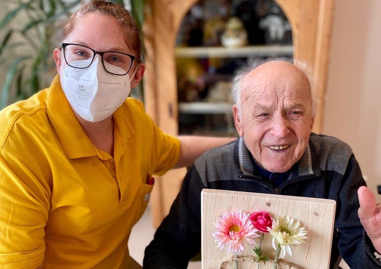 Mitarbeiterin und Bewohner sitzen beieinander. Bewohner hält sein Bild stolz hoch.