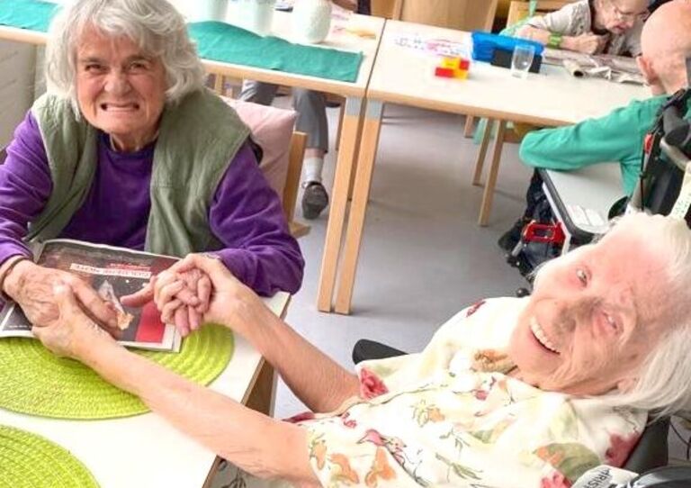 Zwei Bewohnerinnen halten einander am Tisch sitzend an den Händen und blicken fröhlich in die Kamera.