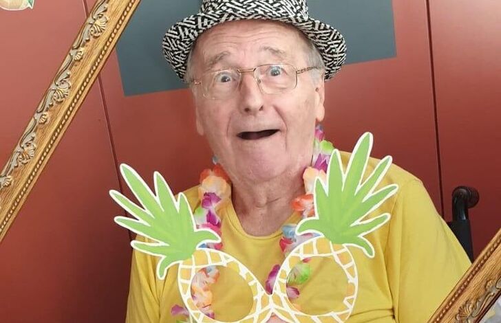 Ein Bewohner mit einer Papierananas am Stiel blickt durch einen leeren Rahmen und macht ein lustiges Gesicht.