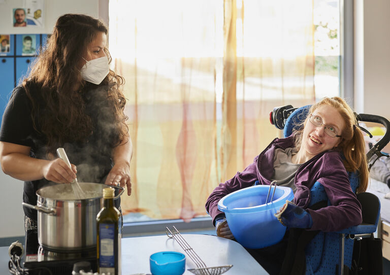 Eine Mitarbeiterin kocht mit Kochtopf und eine Kundin im Rollstuhl sitzt daneben in ihrem Rollstuhl und hält eine Schüssel.