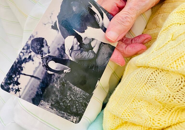 Nahaufnahme einer Hand die eine Scharz-Weiß-Foto in der Hand hält, das sie als Kind abbildet, neben ihr hockt ihre Mutter, die eine Kuh melkt.