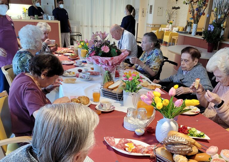 Seitliche Aufnahme vom gut gefüllten Frühstückstisch und en Bewohner*innen, die genüsslich essen.