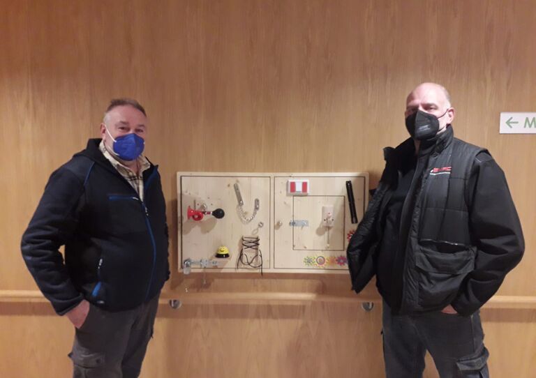 Die zwei ehrenamtlichen Mitarbeiter stehen neben der Wand.