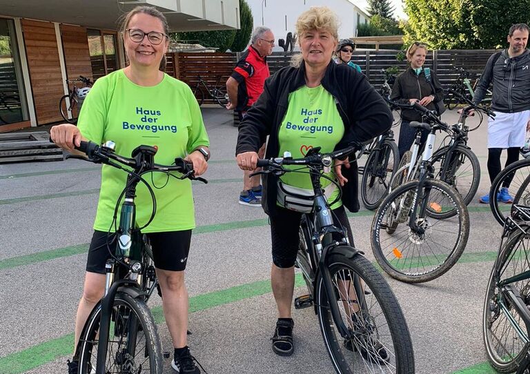 Zwei Mitarbeiter*innen stehen mit ihrem Fahrrad nebeneinander. Sie tragen beide ein T-Shirt "Haus der Bewegung".