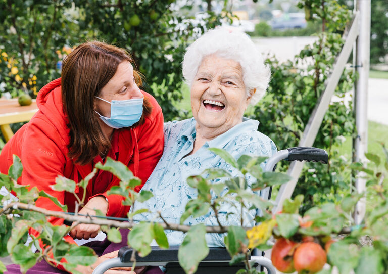 Bewohnerin unter einem Apfelbaum lacht. Mitarbeiterin beugt sich zu ihr.